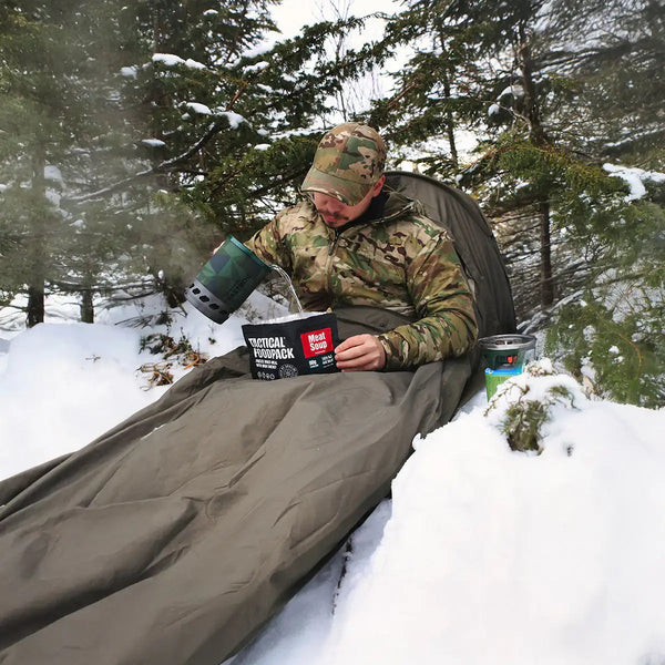 Ein Mann bereitet sich im Schlafsack Notfallnahrung zu, während Schnee um ihn herum liegt.