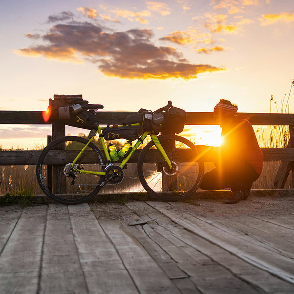 Ein Fahrrad mit einem Mensch daneben stehend auf einer Holzbrücke in der Abenddämmerung.