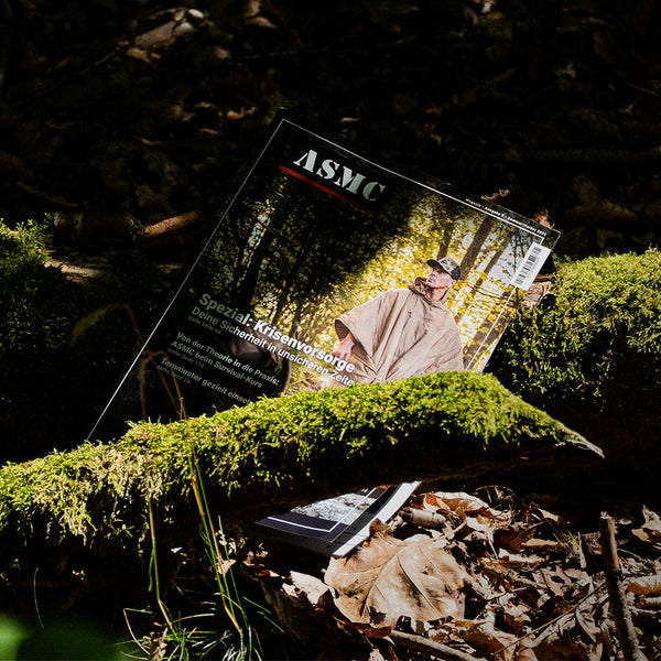 Das ASMC-Magazin auf dem Waldboden mit moosbewachsenen Wurzeln leicht verdeckt.